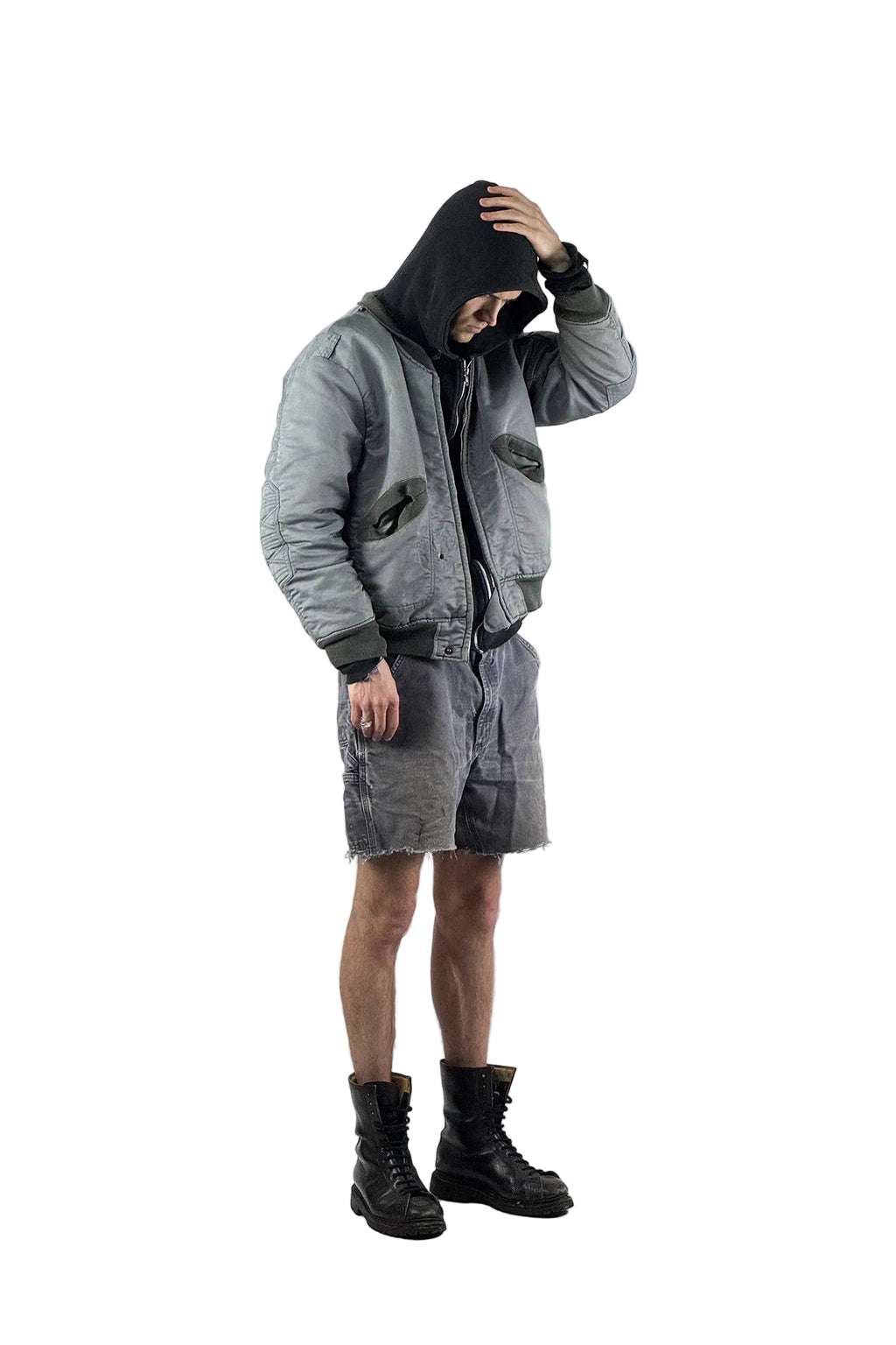 Urbaner Streetwear-Look mit einer grauen Vintage Bomberjacke, schwarzem Hoodie und distressed Carhartt Cut-Off Shorts kombiniert mit schwarzen militärischen Schnürstiefeln vor weißem Hintergrund.