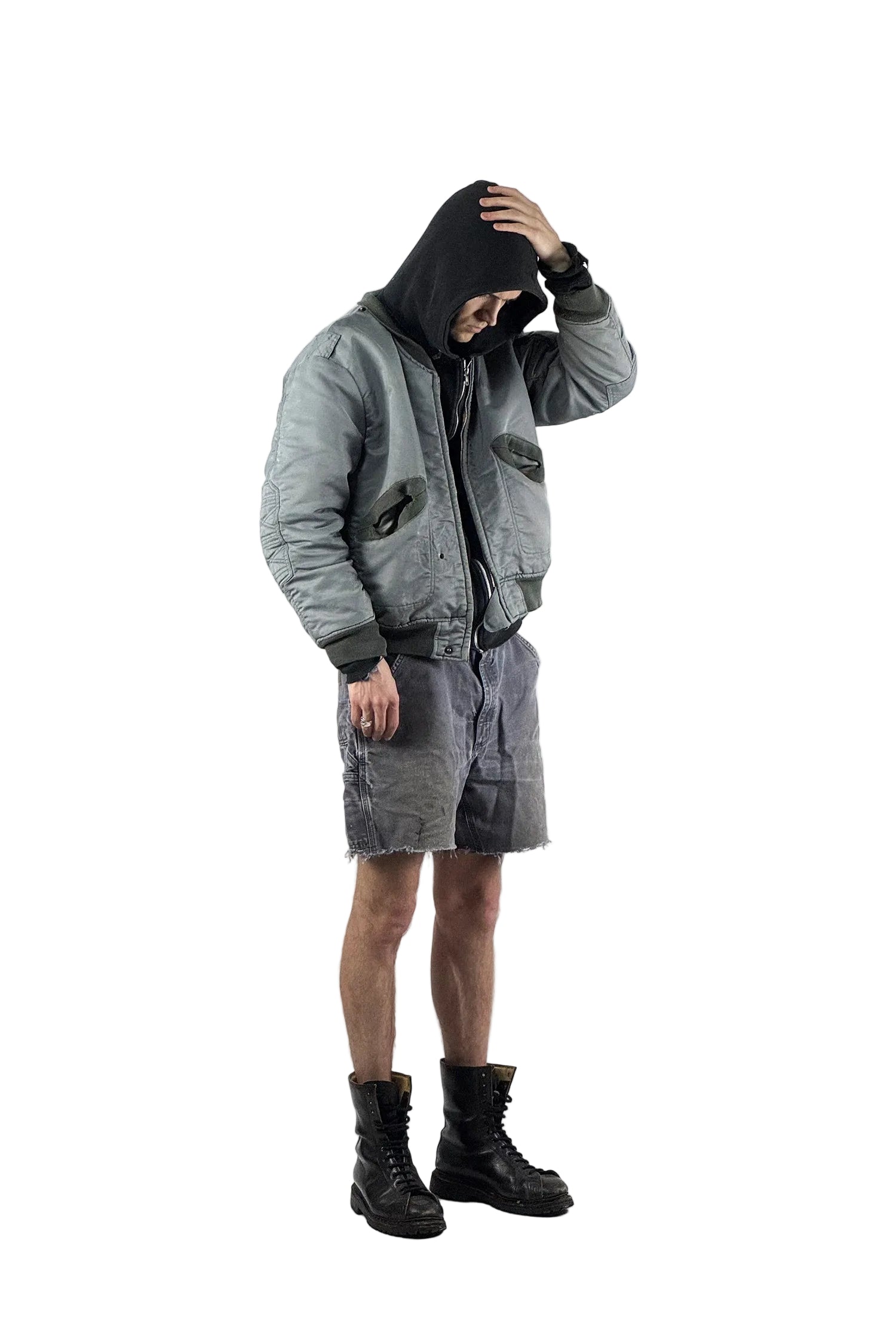 Urbaner Streetwear-Look mit einer grauen Vintage Bomberjacke, schwarzem Hoodie und distressed Carhartt Cut-Off Shorts kombiniert mit schwarzen militärischen Schnürstiefeln vor weißem Hintergrund.