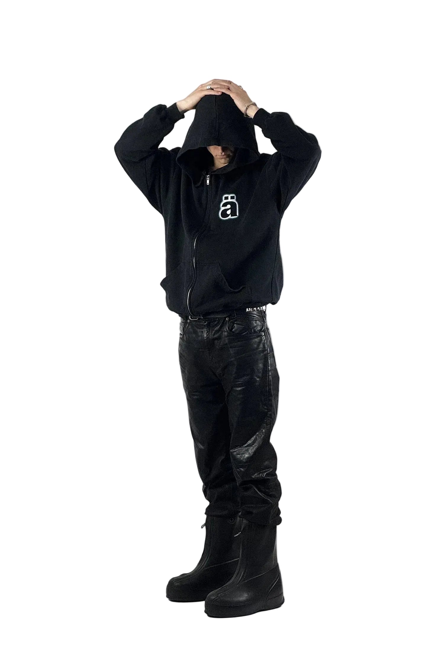 All-Black-Streetwear-Outfit mit einem schwarzen "Die Ärzte" Zip-Up-Hoodie inklusive weißem "Ä" Grafik-Logo, schwarzen Echtlederhosen und klobigen Army Overboots im Balenciaga-Stil vor weißem Studiohintergrund.