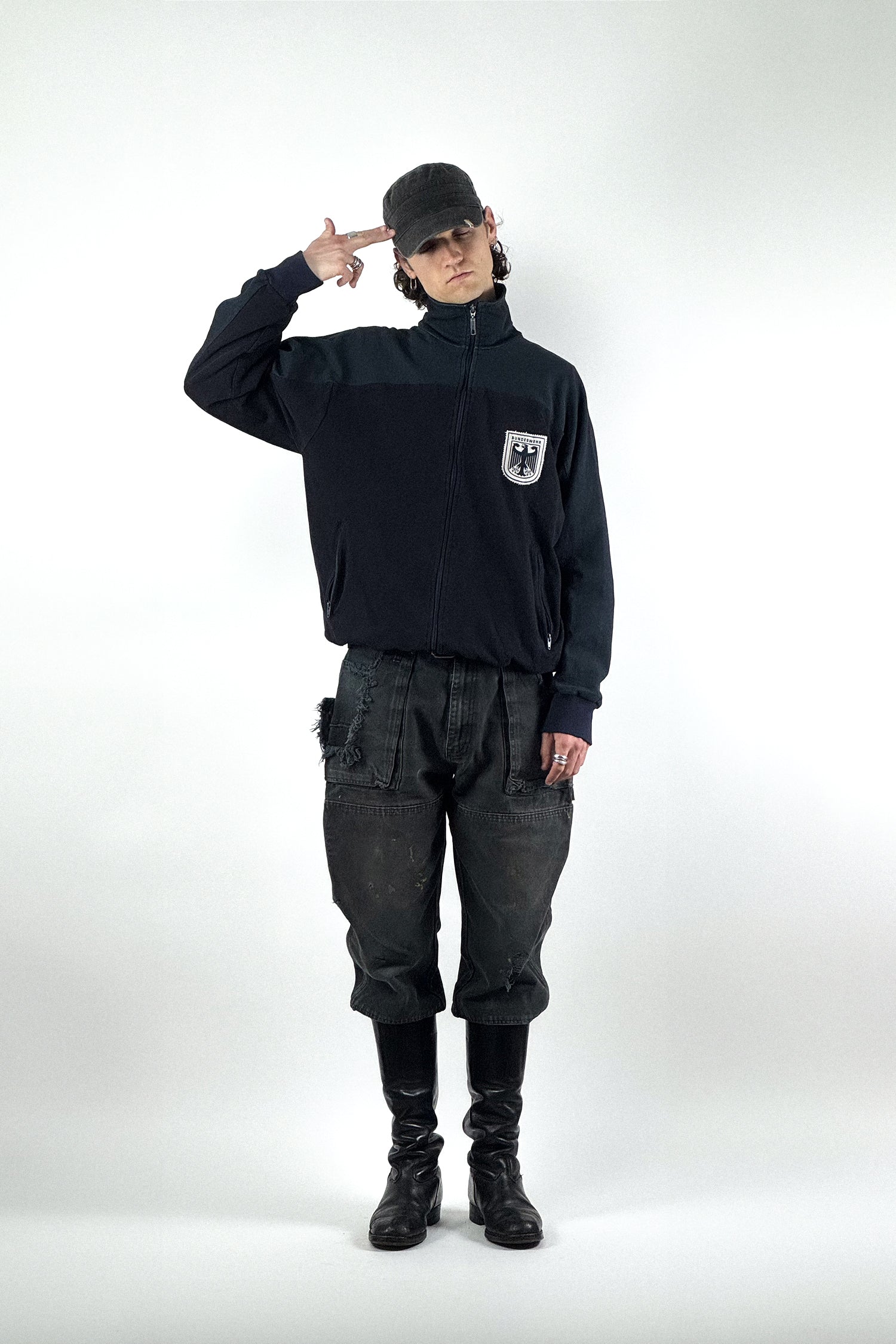 Eine Person steht in einem Studio und zeigt einen Look mit einer originalen Bundeswehr Trainingsjacke mit Adler-Wappen, einer schwarzen Dickies Workwear Hose im Distressed-Look und hohen schwarzen Lederstiefeln.
