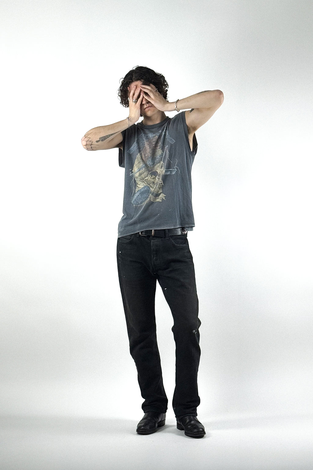 Edgy Streetwear-Outfit mit einem ärmellosen Vintage Grafik-T-Shirt mit Totenkopf-Print, schwarzer Levis 501-Jeans und schwarzen Leder-Biker-Boots vor weißem Hintergrund.