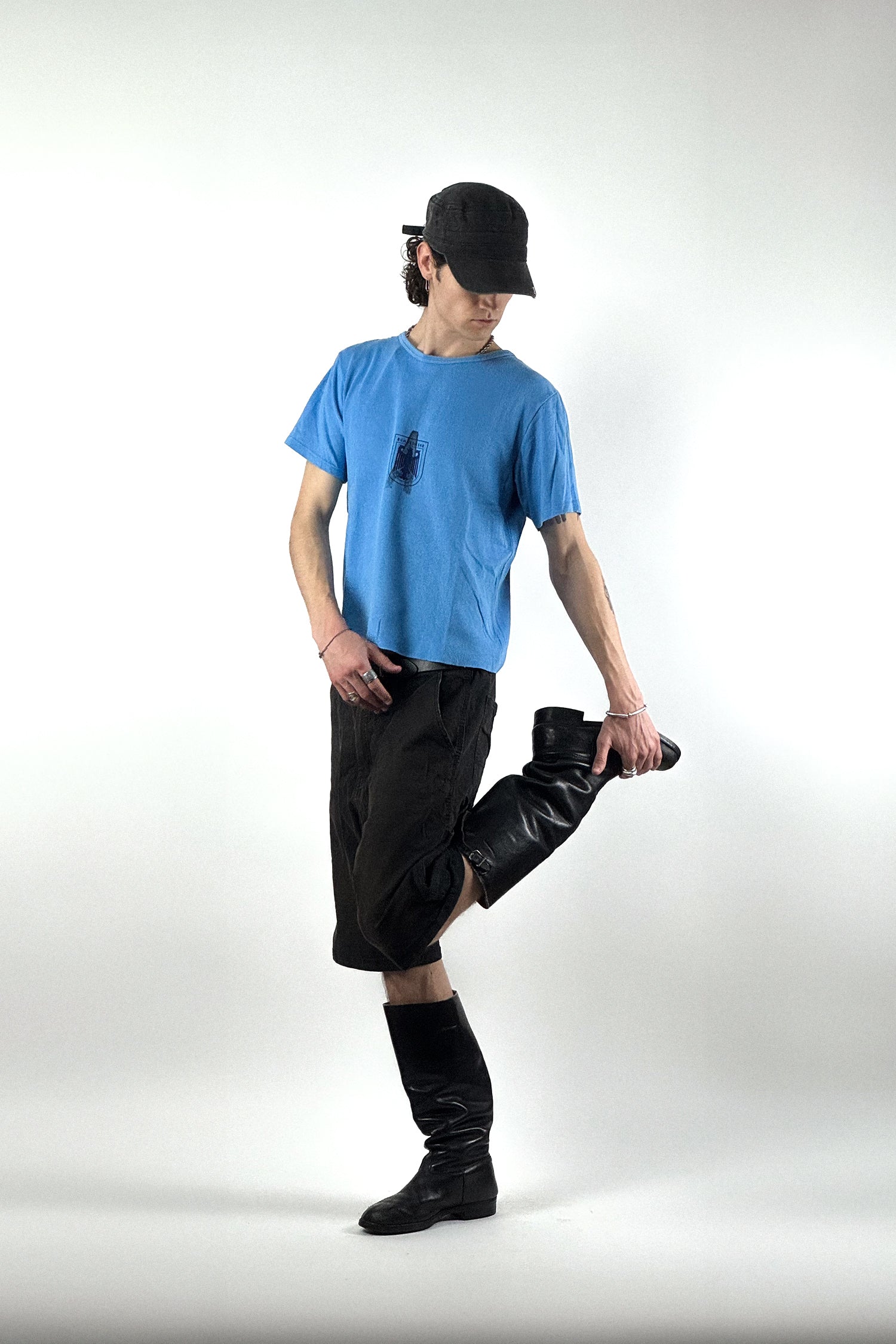 Junger Mann in Vintage Mode vor weißem Hintergrund, trägt ein hellblaues Vintage Bundeswehr T-Shirt mit einem schwarzen Adler-Logo und einem markanten „A“-Stempel darüber, dazu schwarze Shorts und hohe schwarze Lederstiefel.