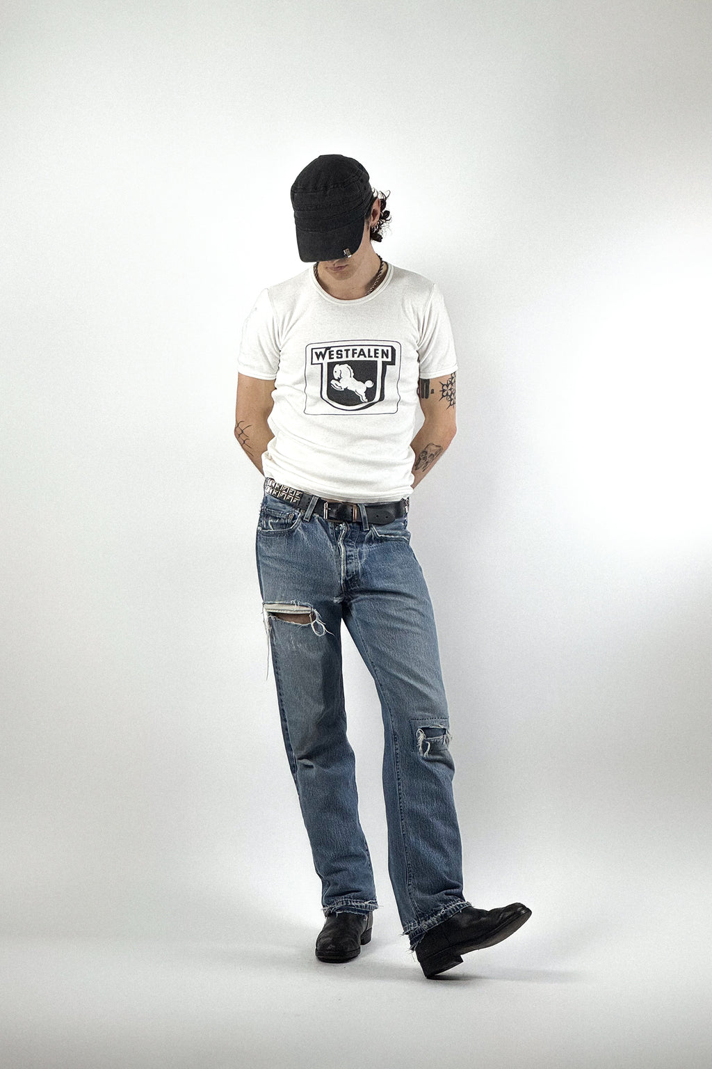 Retro-Streetwear-Outfit mit weißem Vintage Grafik-T-Shirt "Westfalen", blauer Distressed-Jeans mit Rissen am Knie, schwarzer Military-Cap und Lederboots vor weißem Hintergrund.