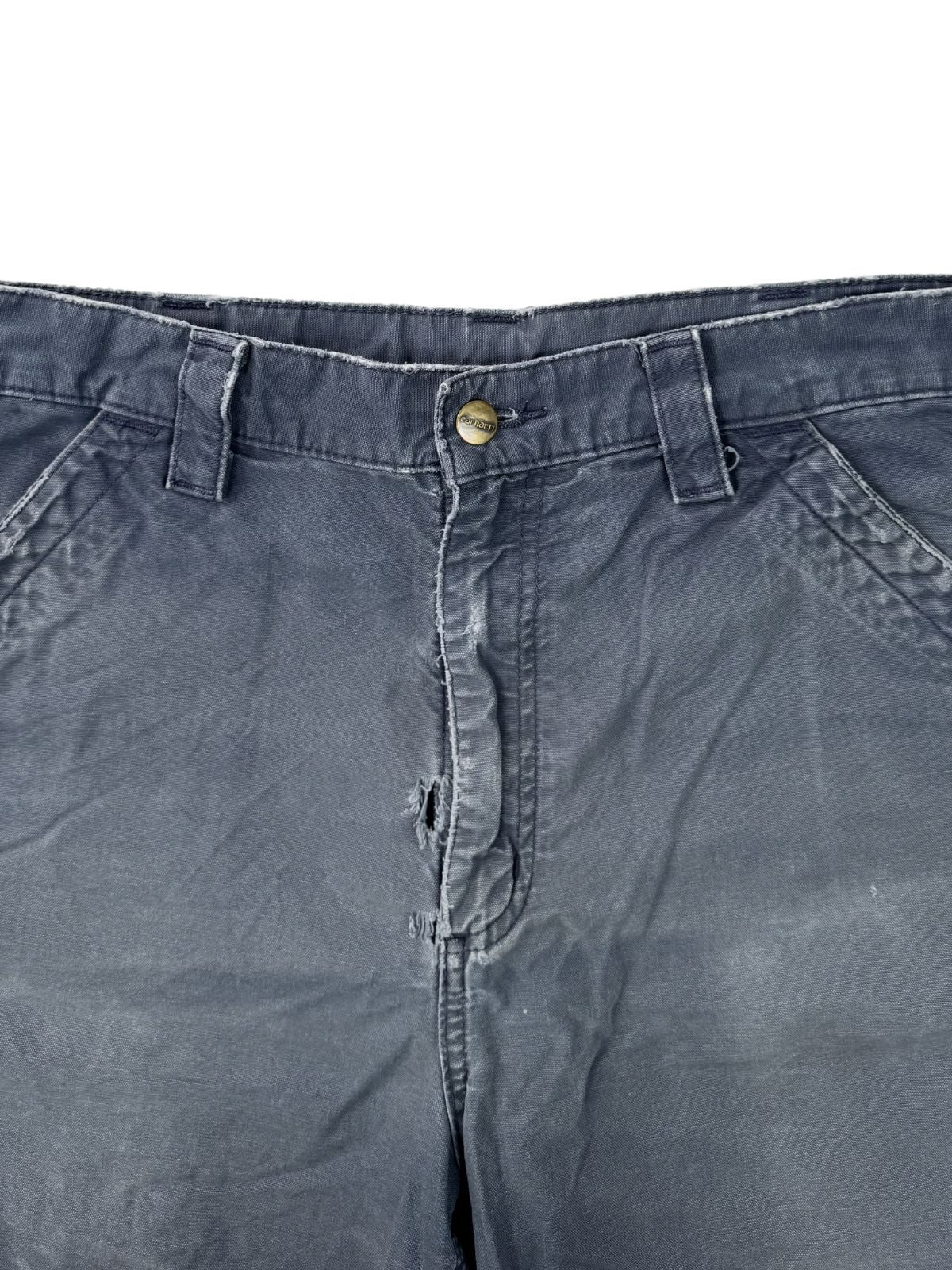Nahaufnahme des metallischen Carhartt-Logoknopfs und des Reißverschlussbereichs einer gebrauchten blauen Arbeitshose.