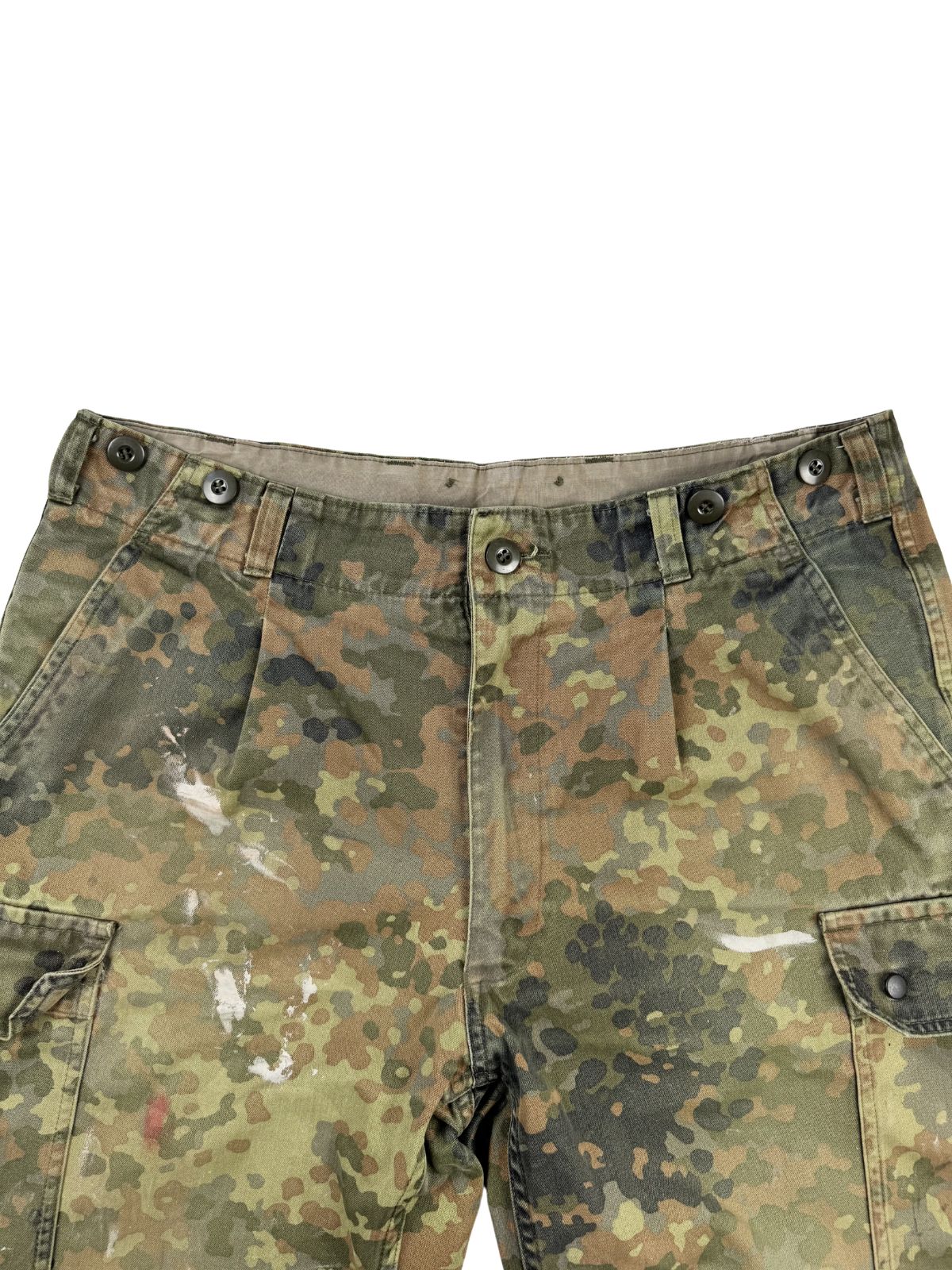 Detailansicht des vorderen Bundes der Camouflage-Shorts mit Knöpfen für Hosenträger, Gürtelschlaufen und dem Hosenbund. Auf dem Flecktarn-Stoff sind deutliche weiße Farbspritzer im Vintage-Stil sichtbar.