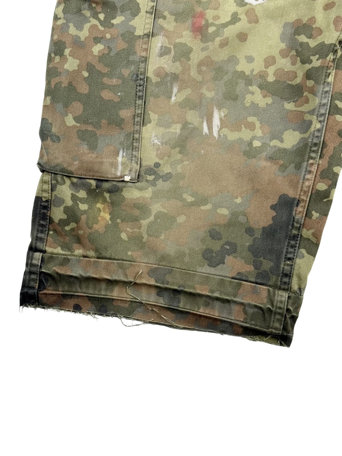 Detailaufnahme des rechten, ungesäumten Hosenbeins zur Veranschaulichung des Used-Looks sowie der weißen Farbspritzer auf dem Camouflage-Gewebe.
