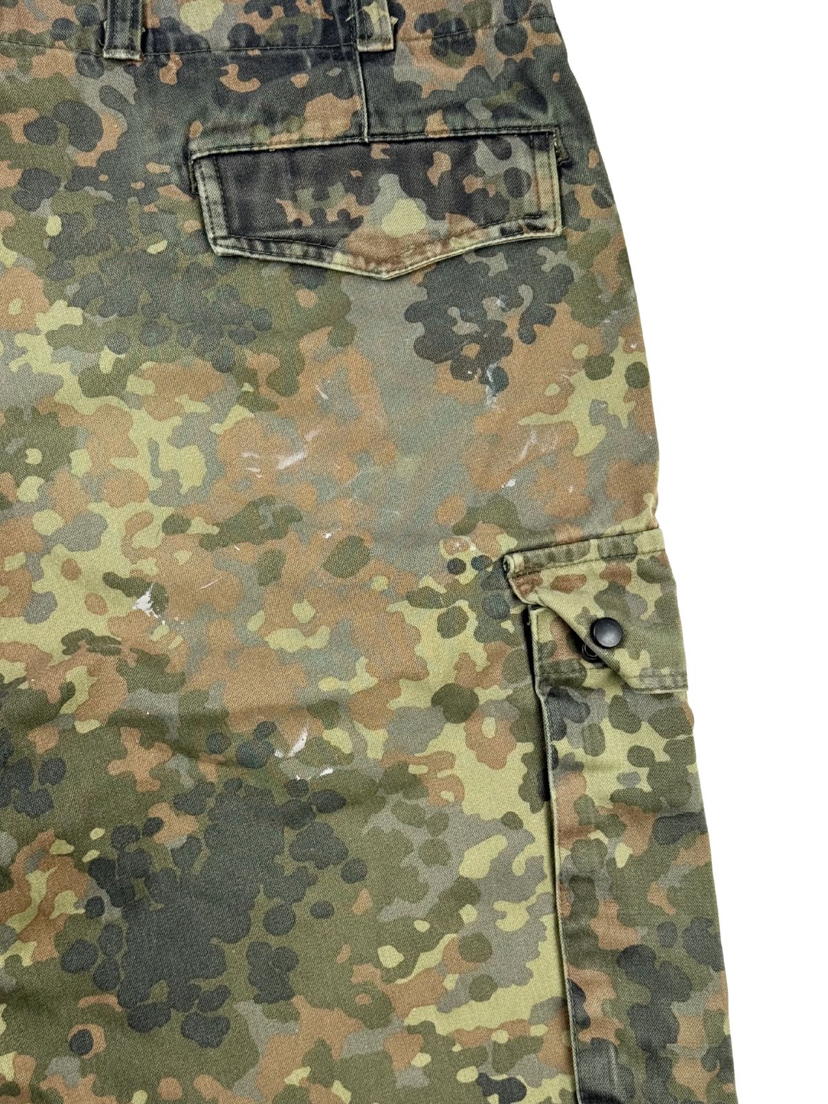 Nahaufnahme der rechten Rückseite zur Zustandsprüfung. Neben der seitlichen Cargo-Tasche mit schwarzem Druckknopf sind mehrere kleine, weiße Farbflecken auf dem Flecktarn-Stoff verteilt.