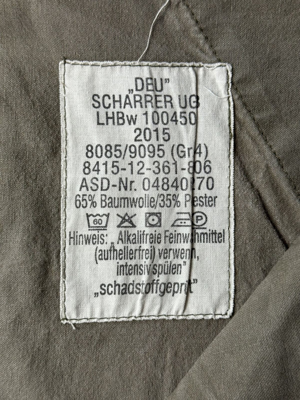 Makroaufnahme des originalen Bundeswehr-Herstelleretiketts auf der Innenseite der Hose. Das weiße Stofflabel enthält offizielle Angaben wie „DEU“ Scharrer UG, das Herstellungsjahr 2015, die Konfektionsgröße Gr. 14, Materialhinweise (65% Baumwolle / 35% Polyester) sowie Pflege- und Waschanleitungen.