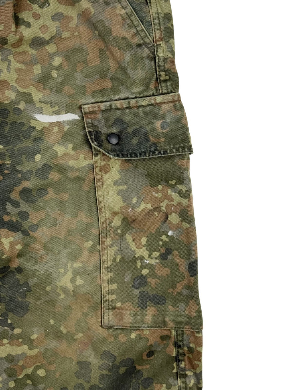Detailansicht der seitlichen Cargo-Beintasche im Flecktarn-Muster. Zu sehen sind die Taschenklappe mit schwarzem Metalldruckknopf sowie charakteristische weiße Farbspritzer auf dem robusten Camouflage-Stoff.