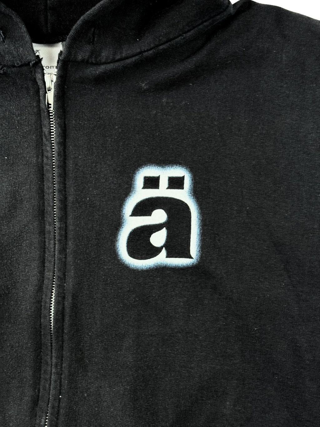Nahaufnahme des Die Ärzte Logos (kleines „ä“ mit Umlautpunkten) in weißem Siebdruck mit blauem Glow-Effekt auf schwarzem Stoff.