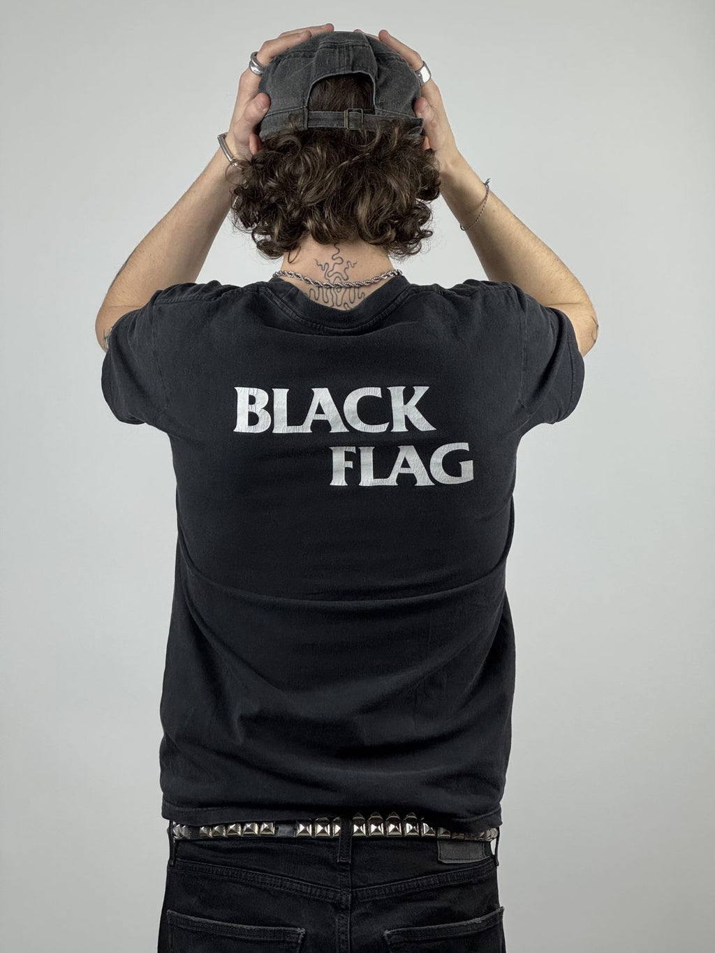 Rückansicht des getragenen Black Flag Shirts kombiniert mit einer schwarzen Mütze; man sieht den weißen „BLACK FLAG“ Schriftzug im Nackenbereich auf dem verwaschenen schwarzen Stoff.