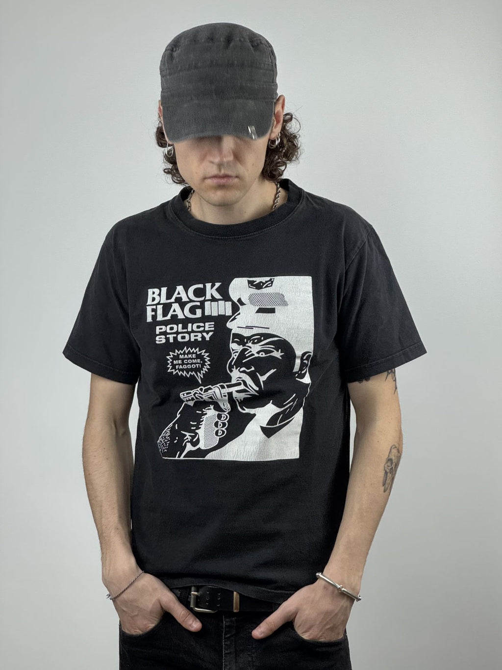 Ein Model mit Mütze trägt das Black Flag „Police Story“ Vintage Shirt; Fokus auf dem provokanten Raymond Pettibon Grafikprint und der lockeren Punk-Rock-Streetwear-Passform.