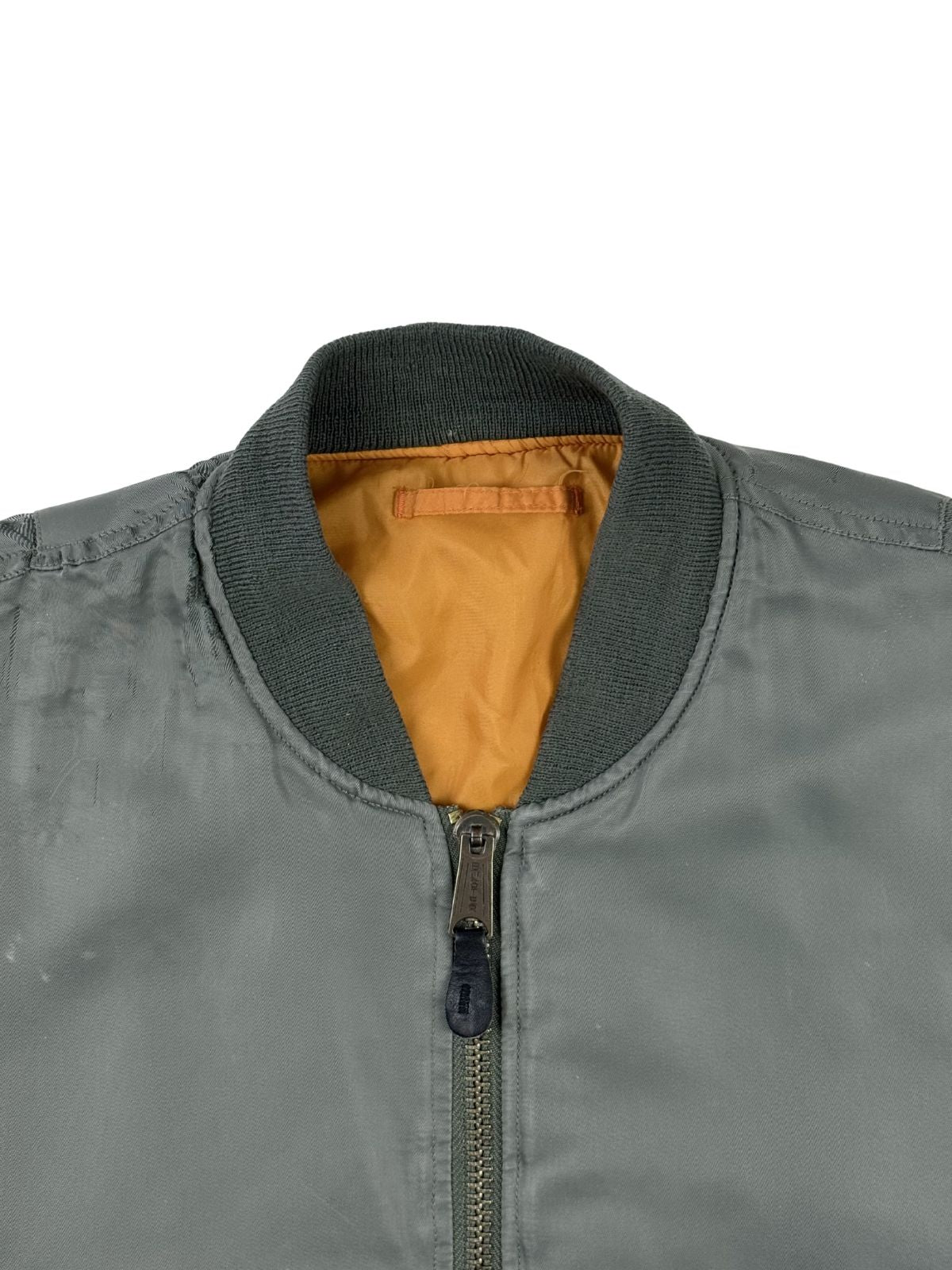 Detailansicht des Kragen- und Reißverschlussbereichs der grauen Bomberjacke. Zu sehen sind das olivgraue Rippstrickbündchen am Hals, der metallene Front-Reißverschluss mit schwarzem Leder-Schiebergriff und das hervorblitzende orangefarbene Futter.