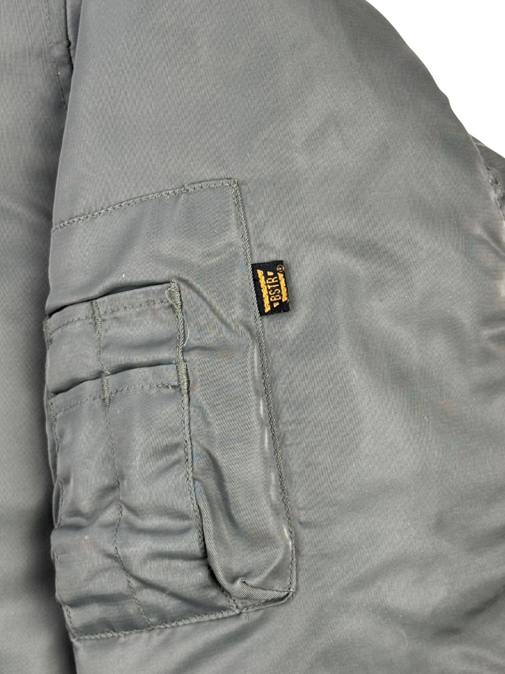 Detailansicht der typischen Utility-Tasche (Ärmeltasche) auf dem linken Oberarm mit Stiftefächern. An der Kante der Reißverschlusstasche ist ein kleines, schwarzes Textillabel mit dem gelben Marken-Schriftzug „BSTR“ (Buster) eingenäht.
