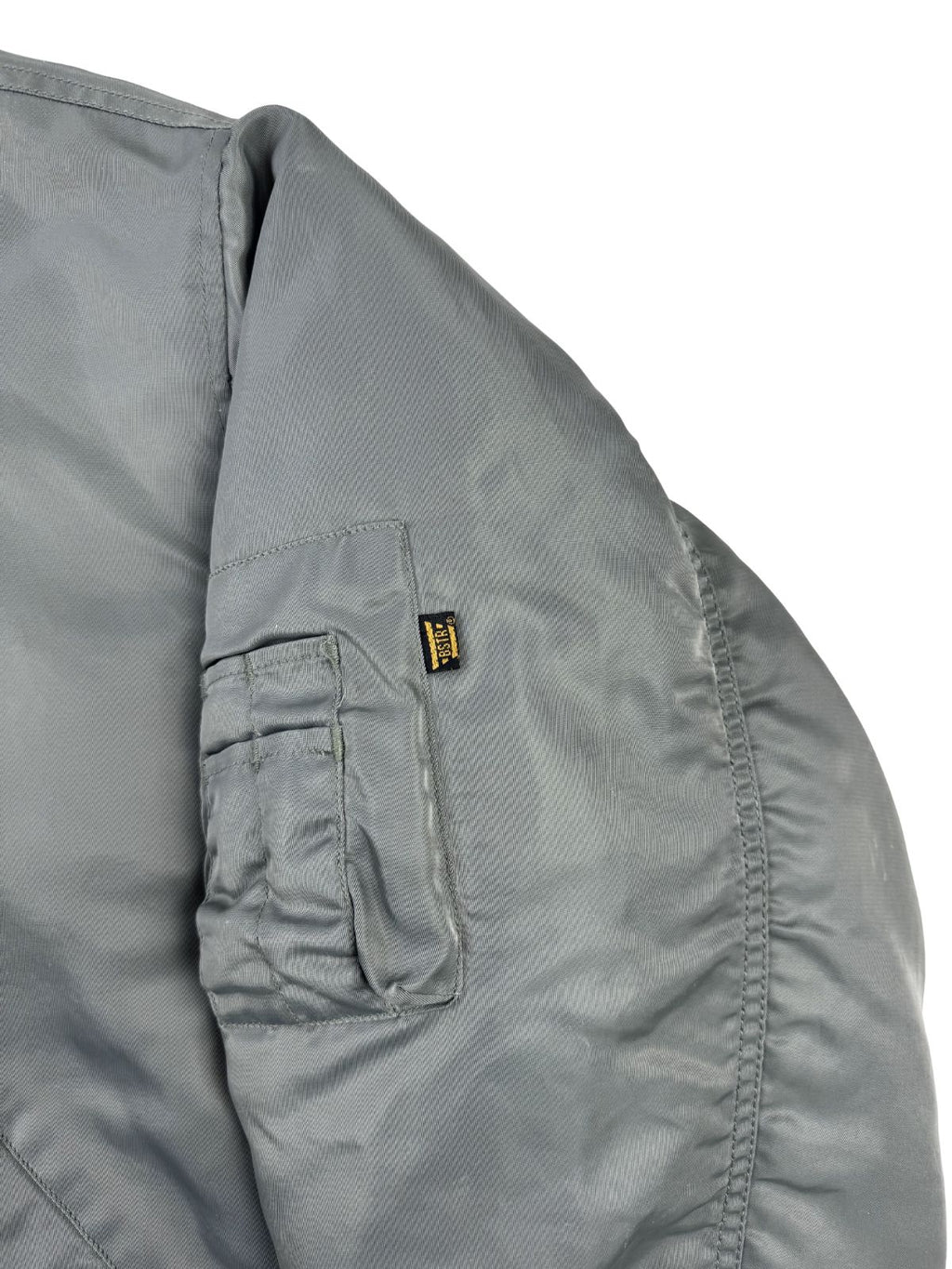 Detailansicht des linken Ärmels der grauen Fliegerjacke. Zu sehen ist die aufgesetzte Utility-Tasche mit Reißverschluss und zwei aufgenähten Stiftefächern sowie das kleine, schwarze Stofflabel der Marke Buster.