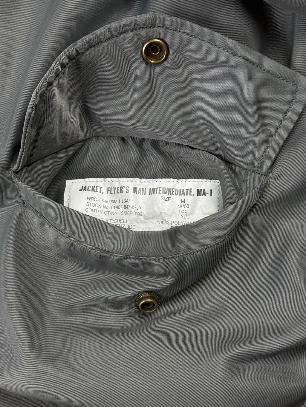 Makroaufnahme des weißen Mil-Spec-Etiketts (Spezifikationsetikett) im Inneren der Seitentasche. Es zeigt offizielle militärische Angaben wie die Modellbezeichnung „JACKET, FLYER'S MAN INTERMEDIATE, MA-1“, die Größe M und Details zur Materialzusammensetzung.