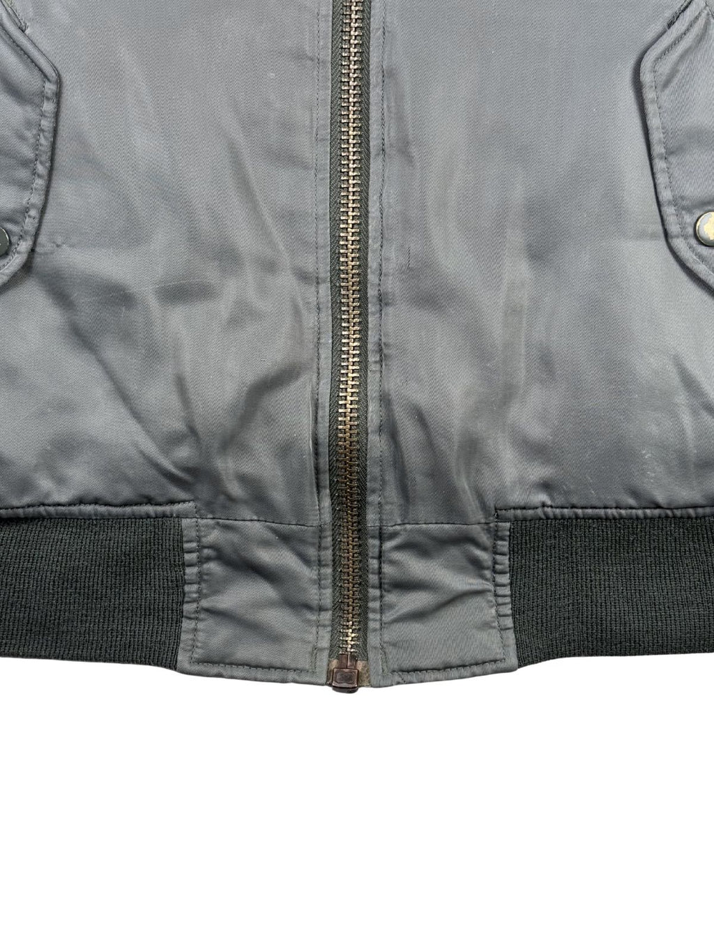 Detailaufnahme des unteren Saumbereichs der Jacke. Zu sehen ist der geschlossene, robuste Metall-Reißverschluss im Übergang zum breiten, elastischen Rippstrickbund in Dunkelgrau.