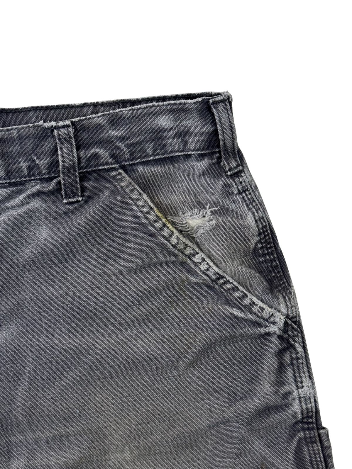 Detailansicht einer vorderen Tasche mit einem Loch und Distressed-Workwear-Look einer grauen Carhartt Shorts.