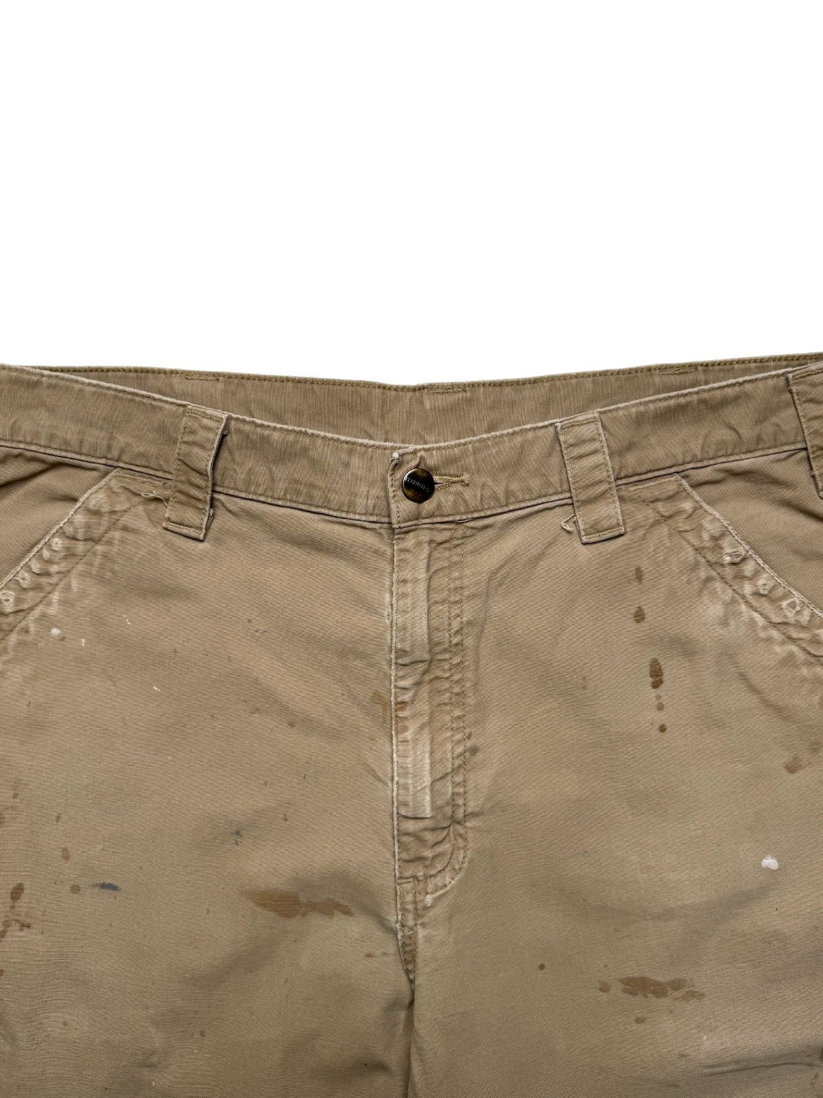Detailaufnahme des oberen vorderen Hosenbunds mit dem klassischen Carhartt Metallknopf, Gürtelschlaufen und den Eingriffstaschen auf distressed Canvas.