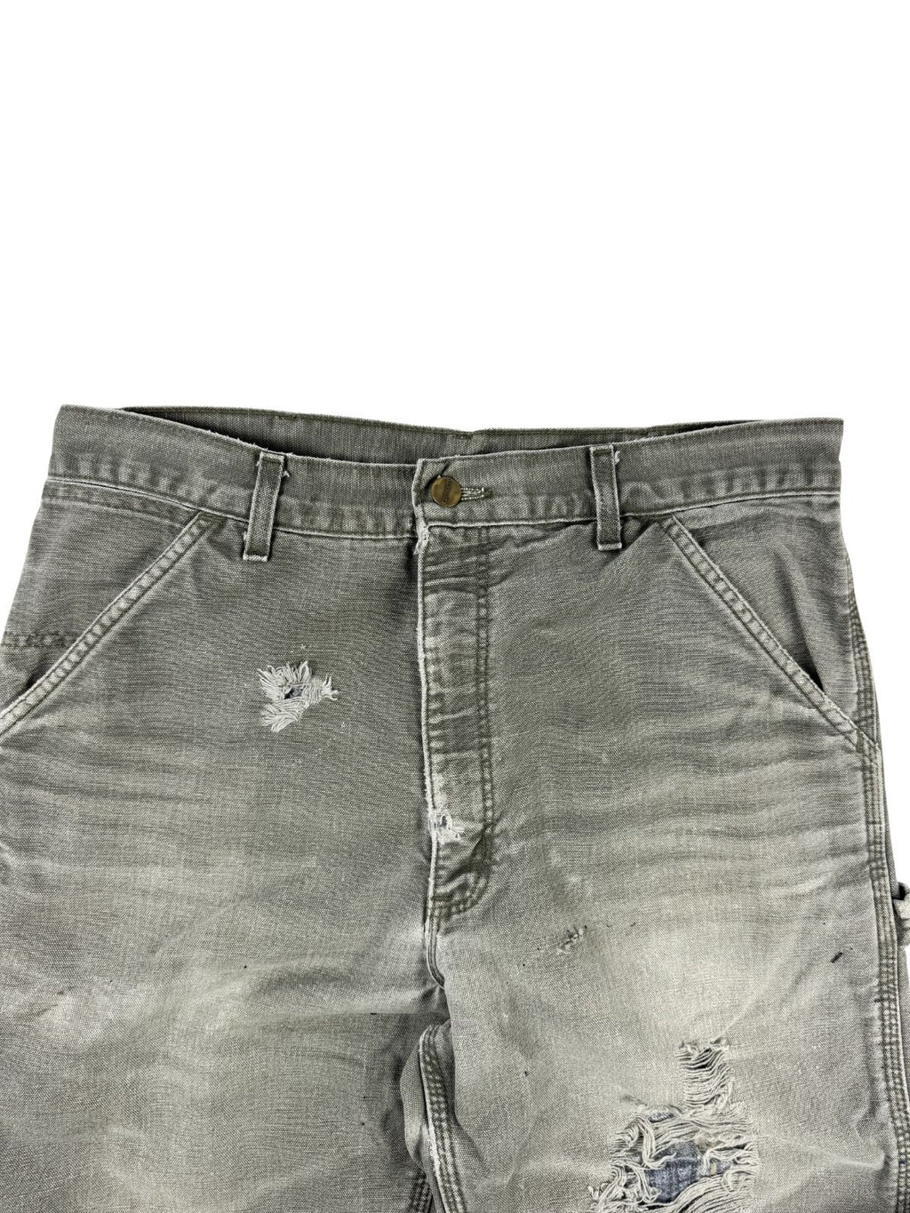 Detailansicht des vorderen Bundes und Schrittbereichs der grauen Carhartt-Hose. Das robuste Canvas-Gewebe zeigt authentische Tragespuren, ein kleineres Loch oberhalb des Oberschenkels und ein großes, ausgefranstes Loch im Kniebereich.