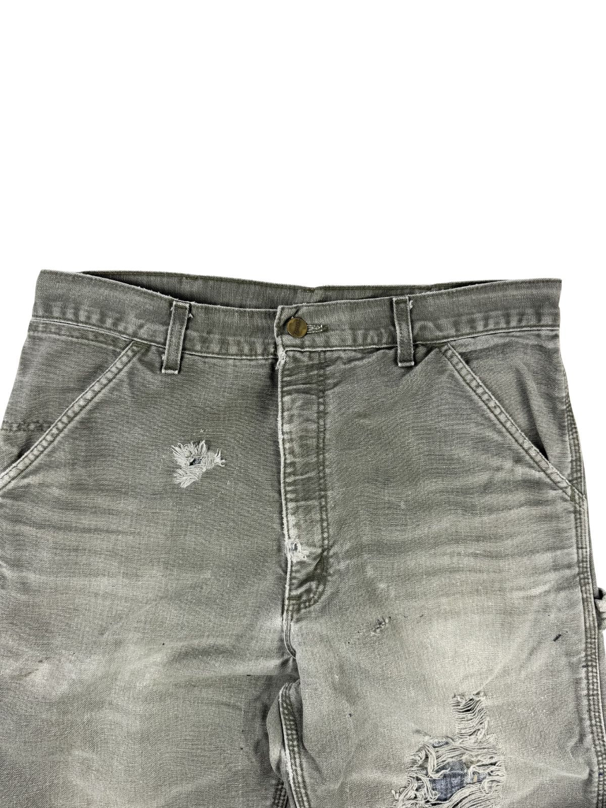 Detailansicht des vorderen Bundes und Schrittbereichs der grauen Carhartt-Hose. Das robuste Canvas-Gewebe zeigt authentische Tragespuren, ein kleineres Loch oberhalb des Oberschenkels und ein großes, ausgefranstes Loch im Kniebereich.