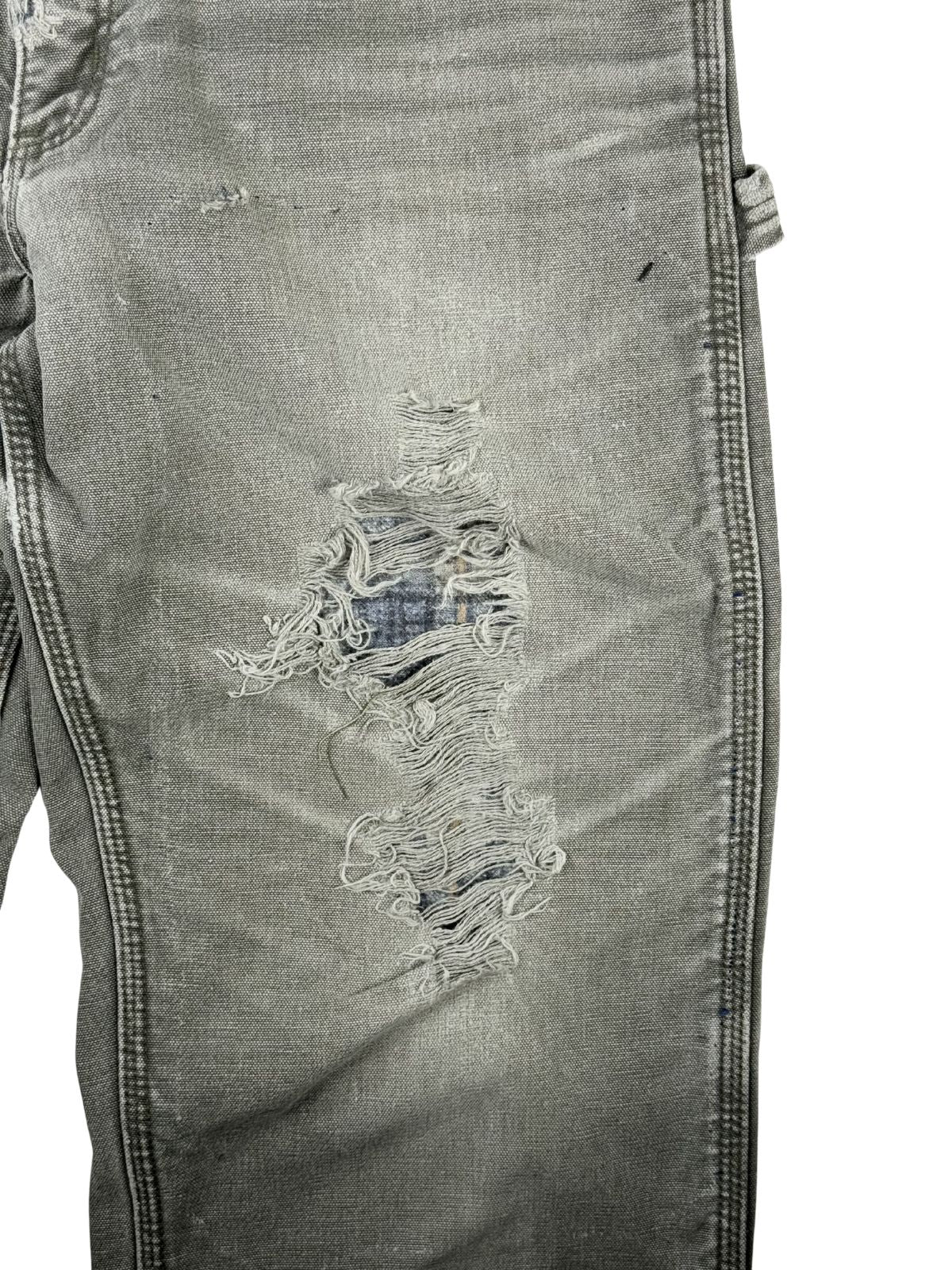 Detailaufnahme des linken Kniebereichs mit einem großen, thrashed Loch. Durch die zerschlissenen, weißen Fäden des Oberstoffs kommt das darunter liegende, karierte Flanellfutter zum Vorschein.