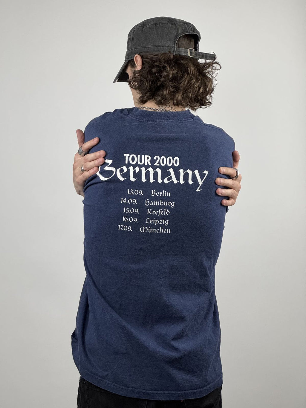 Person trägt das dunkelblaue Cockney Rejects Tour-Shirt von 2000; Fokus auf dem weißen Backprint mit den Tourdaten und der lockeren Vintage-Passform.