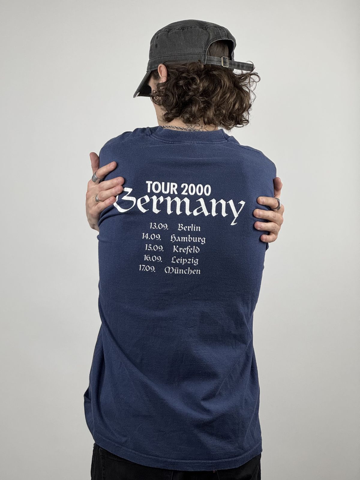 Person trägt das dunkelblaue Cockney Rejects Tour-Shirt von 2000; Fokus auf dem weißen Backprint mit den Tourdaten und der lockeren Vintage-Passform.