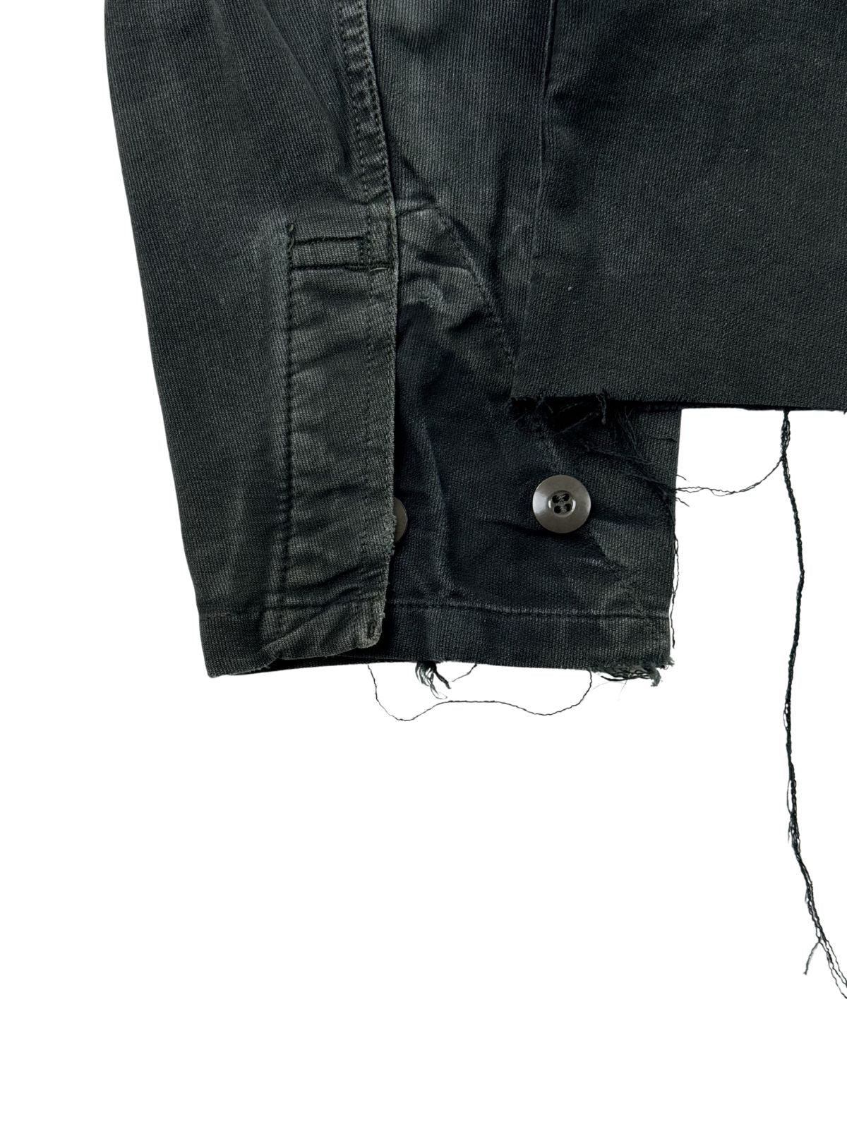 Detailaufnahme des linken Ärmelbündchens mit verstellbarem Knopfverschluss, die zudem die offene, ausgefranste Kante des kurzen Jackensaums zeigt.