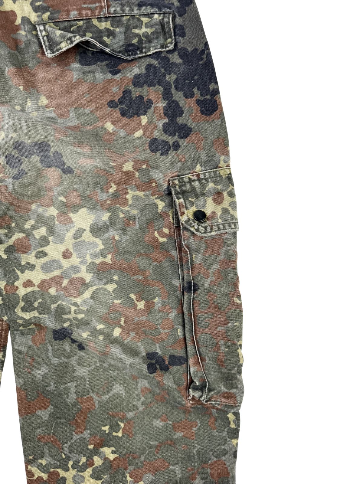 Detailaufnahme der seitlichen, aufgesetzten Cargo-Beintasche im Flecktarn-Muster, verschließbar mit einem verdeckten Druckknopf.