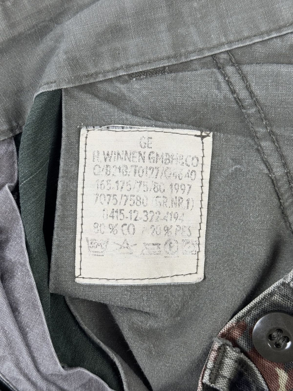 Makroaufnahme des originalen Bundeswehr-Etiketts der Firma „H. Winnen GmbH & Co“. Es zeigt historische Angaben wie das Herstellungsjahr 1997, die Militärgröße „165-175/75/80 (Gr.Nr. 1)“ sowie die Materialzusammensetzung „80% CO / 20% PES“ (80% Baumwolle, 20% Polyester).