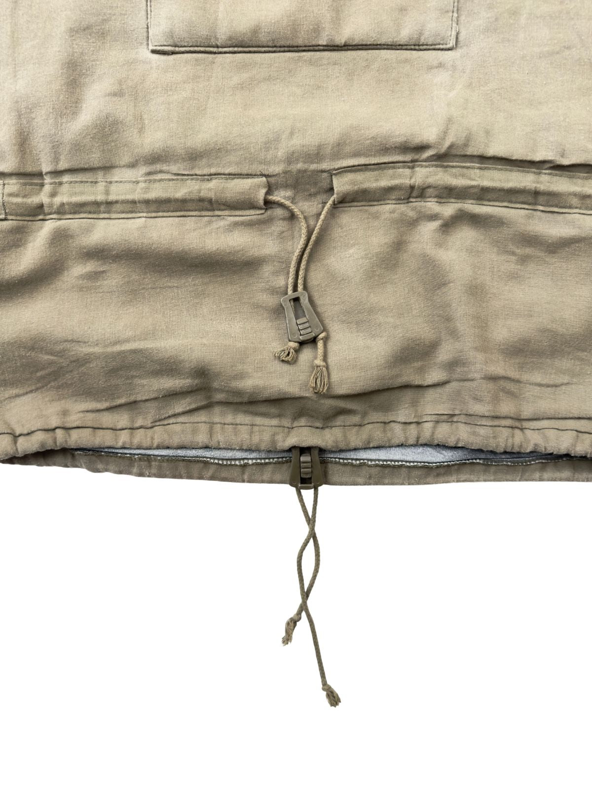 Detailansicht des unteren Bereichs des olivgrünen Vintage Bundeswehr Smocks. Zu sehen sind der verstellbare Kordelzug im Taillentunnel mit einem olivfarbenen Kunststoff-Stopper sowie ein weiterer Kordelzug direkt am unteren Saum des Anoraks.