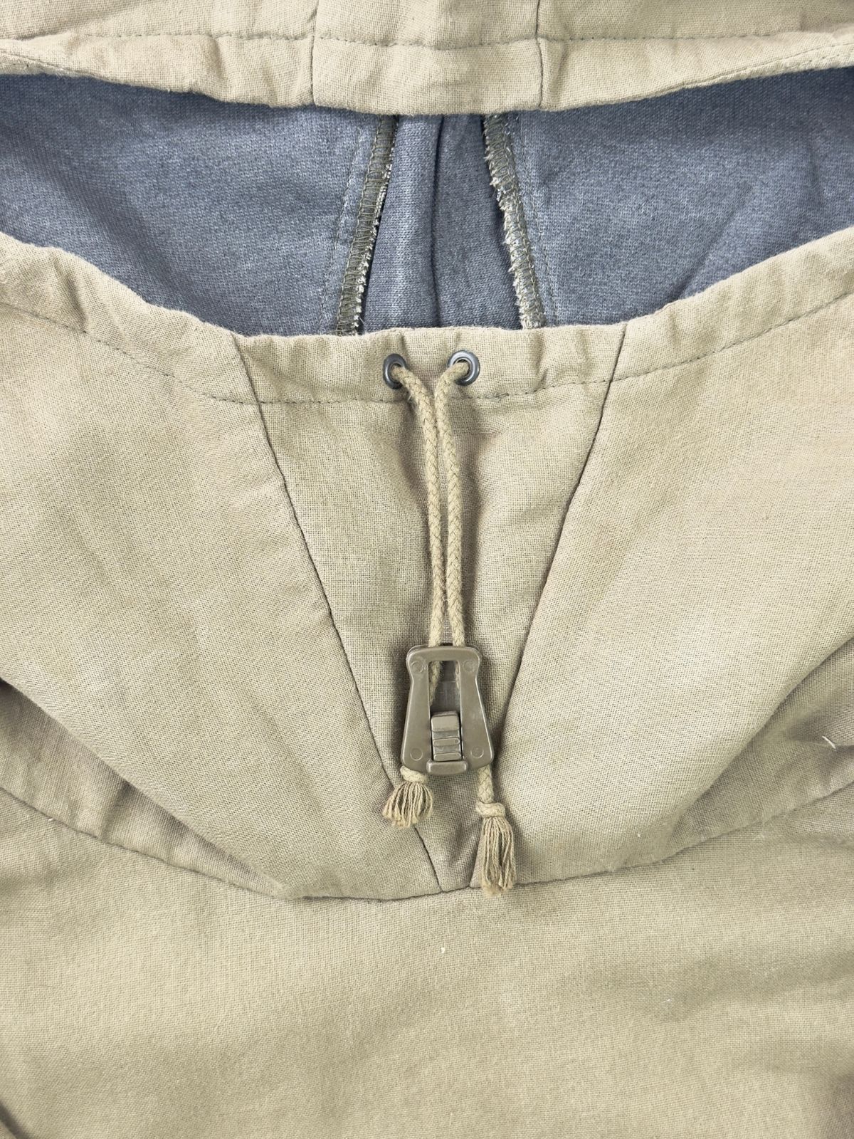Makroaufnahme des Kapuzenverschlusses des Vintage-Smocks mit Fokus auf die Kordelführung durch Metallösen und den olivfarbenen Kordelstopper aus Kunststoff.