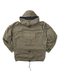 Komplettansicht der Vorderseite eines olivgrünen Vintage Bundeswehr Militär-Smocks aus den 1980er Jahren. Der Schlupfanorak (Windbreaker) verfügt über eine große Kängurutasche auf der Brust, Kordelzüge an Taille und Saum sowie Deutschland-Patches auf den Ärmeln. Flatlay auf weißem Hintergrund.