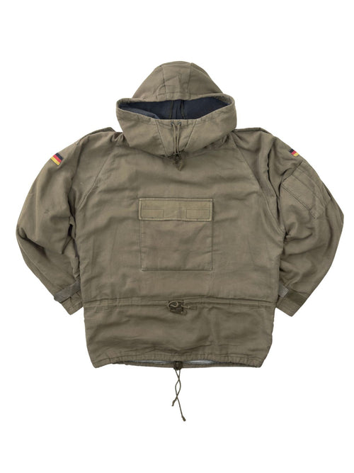 Komplettansicht der Vorderseite eines olivgrünen Vintage Bundeswehr Militär-Smocks aus den 1980er Jahren. Der Schlupfanorak (Windbreaker) verfügt über eine große Kängurutasche auf der Brust, Kordelzüge an Taille und Saum sowie Deutschland-Patches auf den Ärmeln. Flatlay auf weißem Hintergrund.