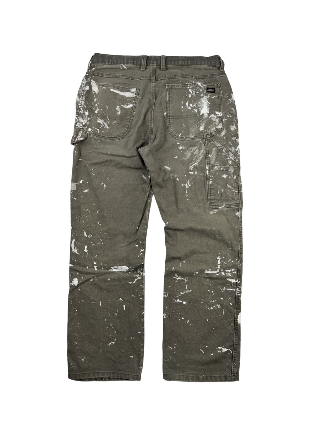Komplettansicht der Rückseite einer oliv-braunen Vintage Dickies Maler-Arbeitshose (Dungaree Pants) im Flatlay. Die Workwear-Hose weist am gesamten Gesäß und den Beinen markante weiße Farbkleckse im echten Painter-Look auf. Flatlay auf weißem Hintergrund.