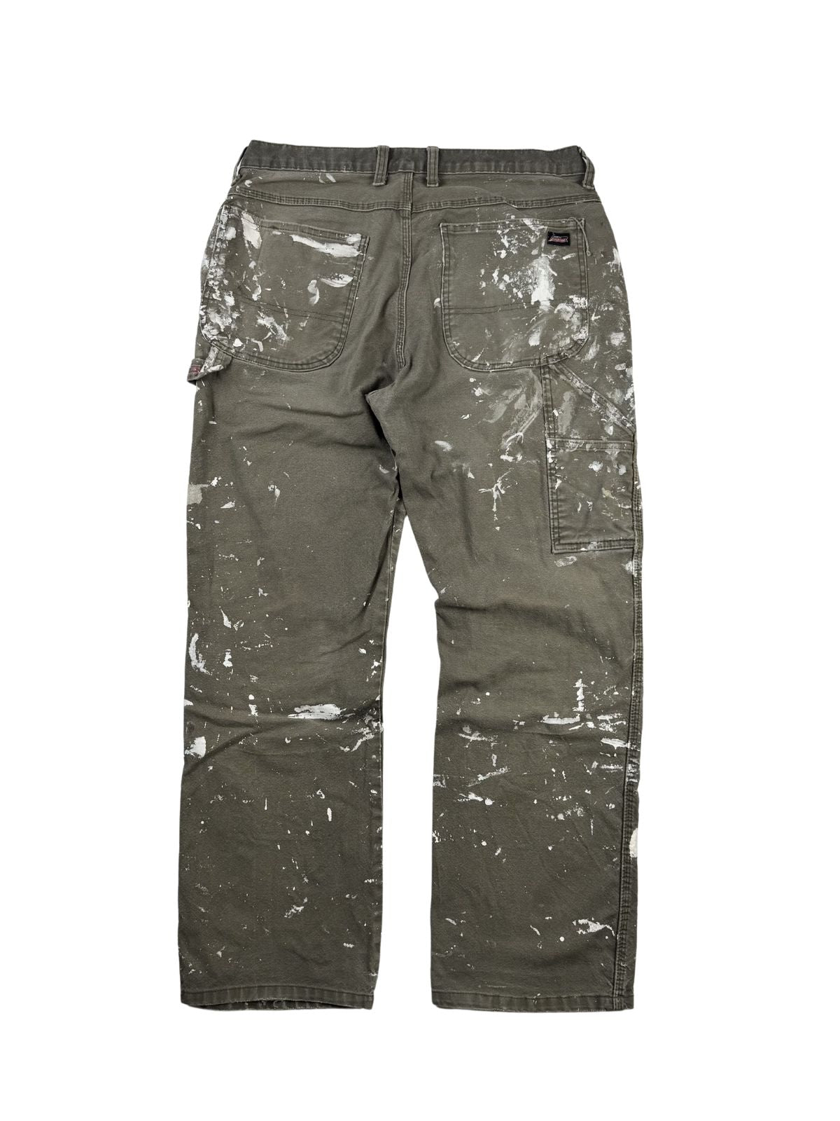 Komplettansicht der Rückseite einer oliv-braunen Vintage Dickies Maler-Arbeitshose (Dungaree Pants) im Flatlay. Die Workwear-Hose weist am gesamten Gesäß und den Beinen markante weiße Farbkleckse im echten Painter-Look auf. Flatlay auf weißem Hintergrund.