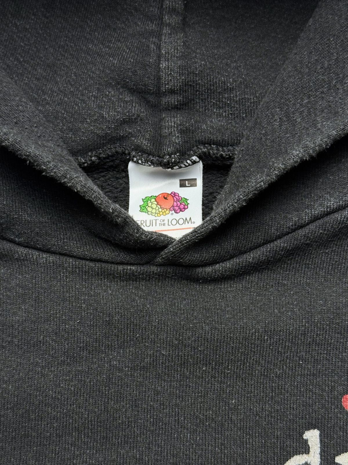Nahaufnahme der Kapuzenbasis mit dem originalen weißen „Fruit of the Loom“-Nackenetikett in Größe L auf verwaschenem schwarzen Sweatstoff.