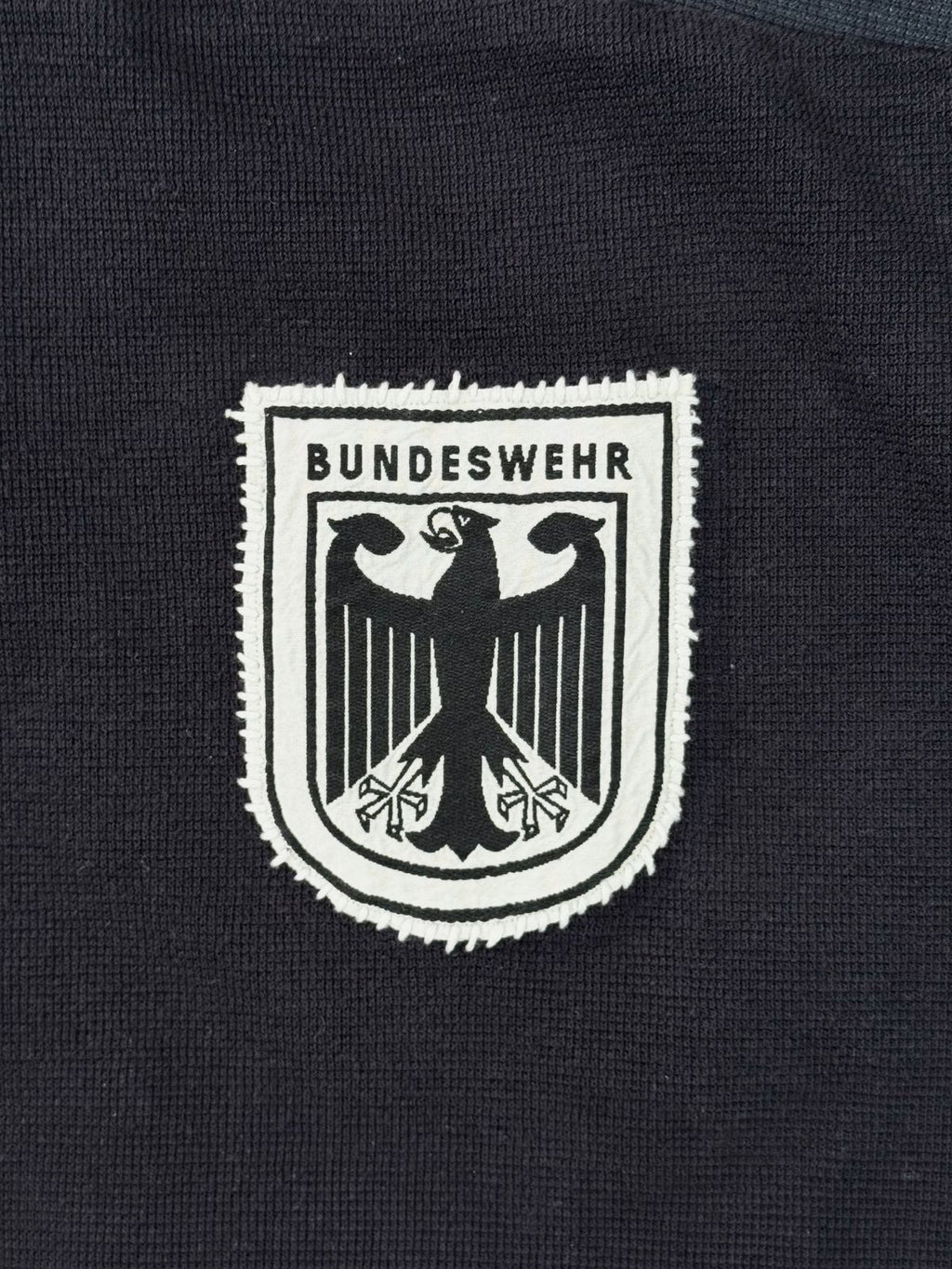 Makroaufnahme des handgestickten Bundeswehr-Wappens; weißes Patch mit schwarzem Adler-Stickmotiv auf dunkelblauem Strickstoff.