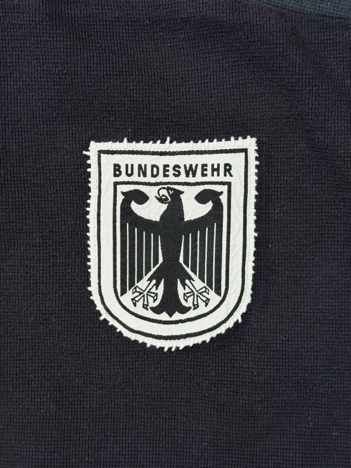 Makroaufnahme des handgestickten Bundeswehr-Wappens; weißes Patch mit schwarzem Adler-Stickmotiv auf dunkelblauem Strickstoff.