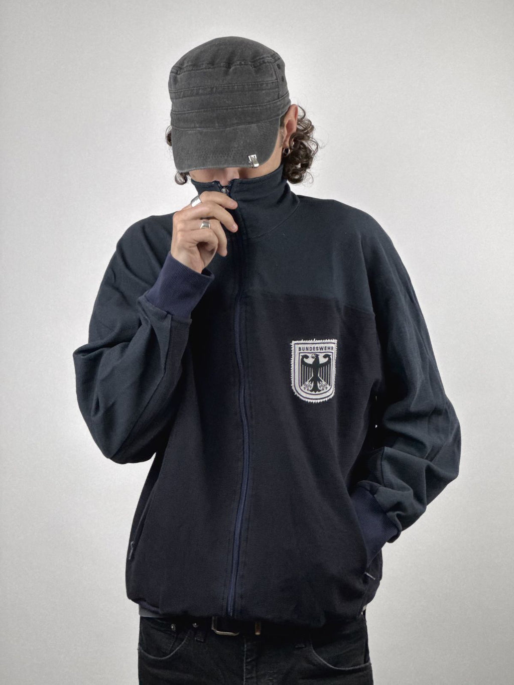Eine Person trägt die eingefärbte Bundeswehr Jacke; Fokus auf dem hochgezogenen Reißverschluss und der markanten Logo-Stickerei.