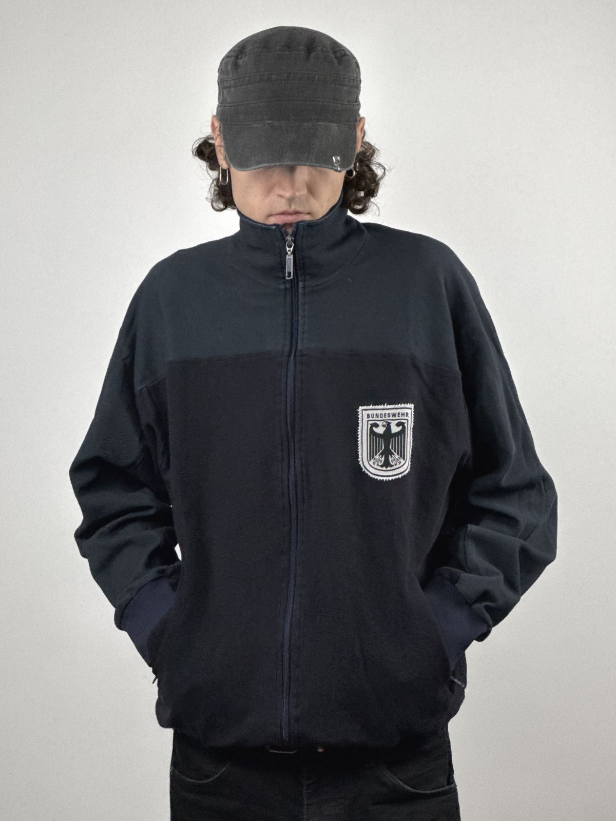 Frontalansicht der Vintage Bundeswehr Jacke im Streetwear-Look; kombiniert mit einer schwarzen Cap und Händen in den Taschen.