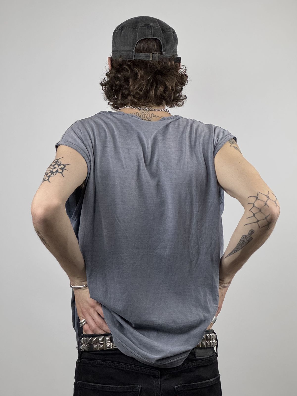 Rückansicht des Modells im ärmellosen Shirt; verdeutlicht den weich fallenden Stoff und die Kombination mit einem Punk-Style Nietengürtel.