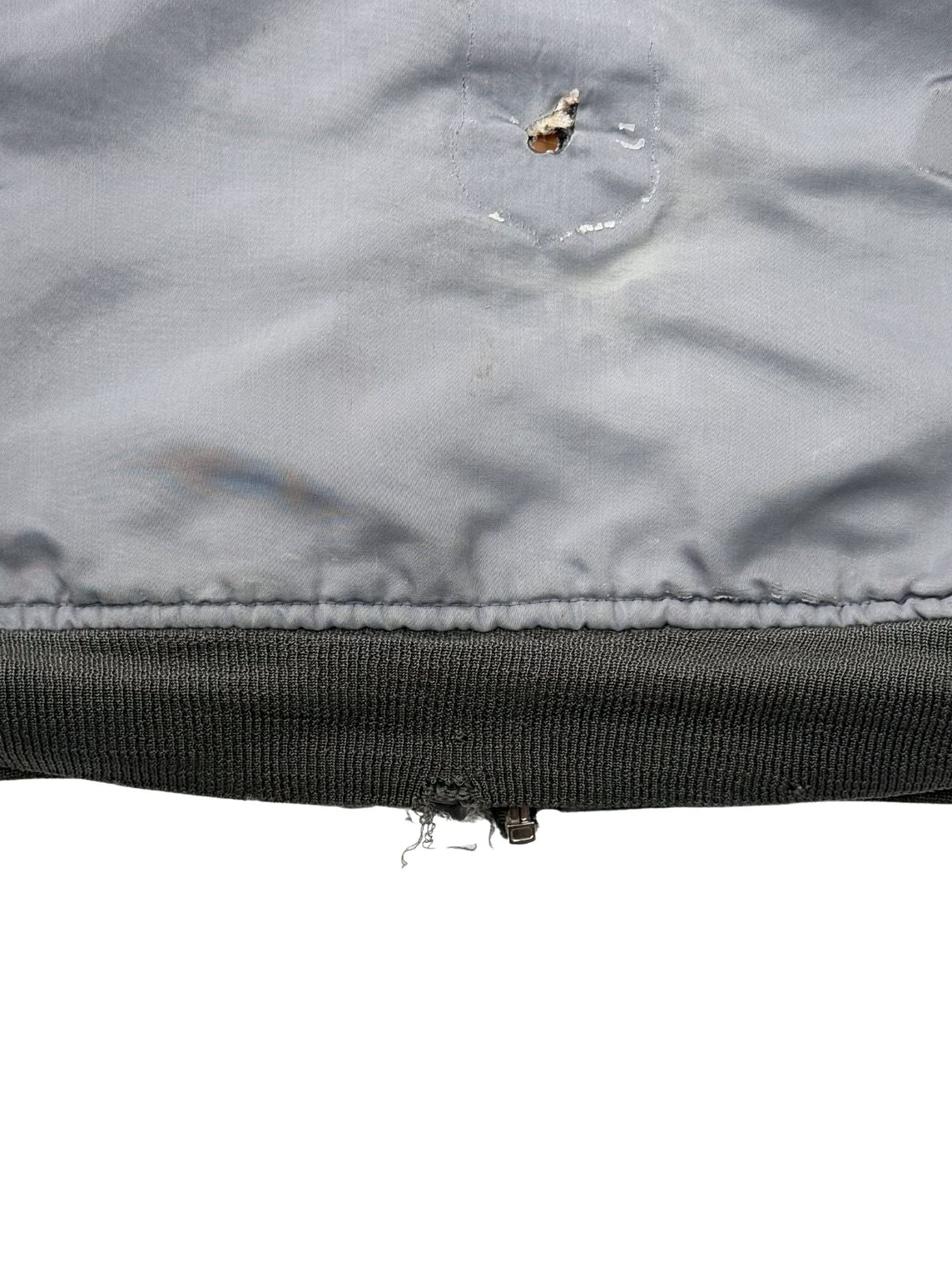 Detailansicht des unteren Jackenrandes; zeigt eine ausgefranste Stelle im Rippstrick-Bündchen und ein markantes Brandloch im hellblauen Obermaterial.