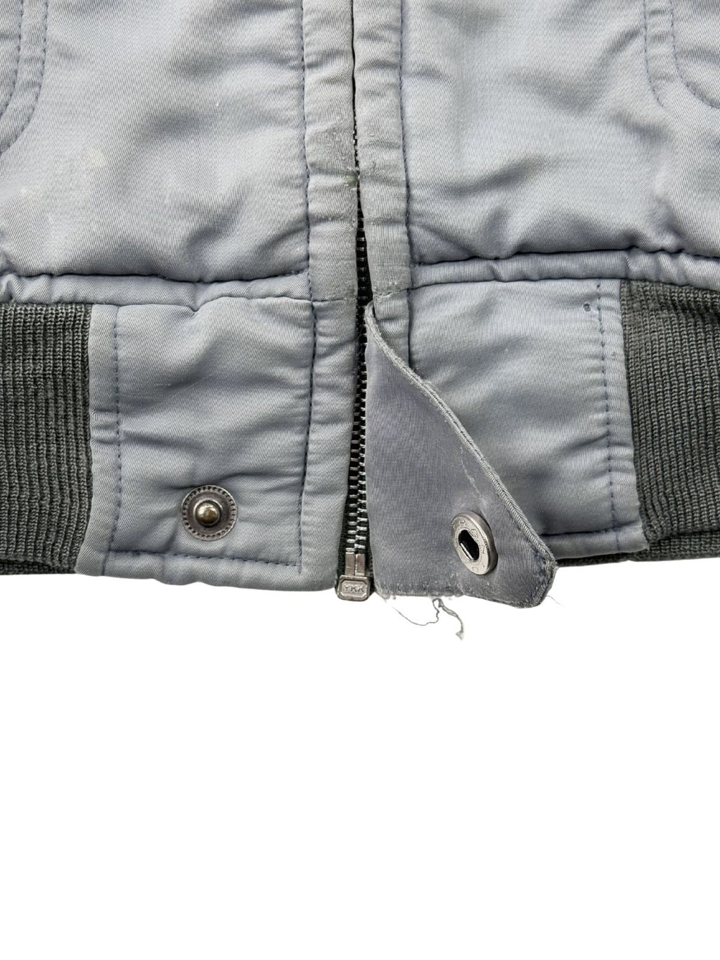 Nahaufnahme des unteren Saums der hellblauen Bomberjacke; zeigt den massiven YKK-Metallreißverschluss, die Rippstrickbündchen und einen funktionstüchtigen Druckknopf am verwaschenen Bund.