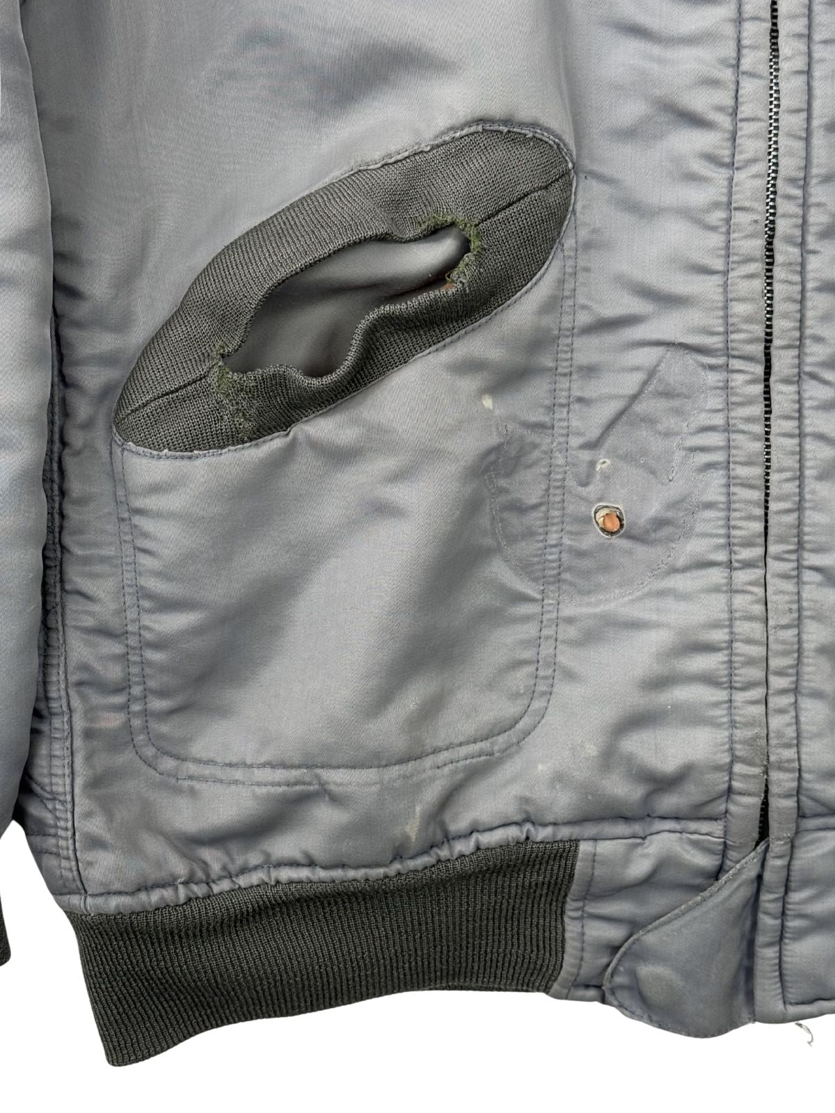 Makroaufnahme des Tascheneingriffs; zeigt starke Abnutzungen (Fraying) am grünen Rippstrick-Saum und ein kleines Brandloch auf der Frontseite der vintage Fliegerjacke.
