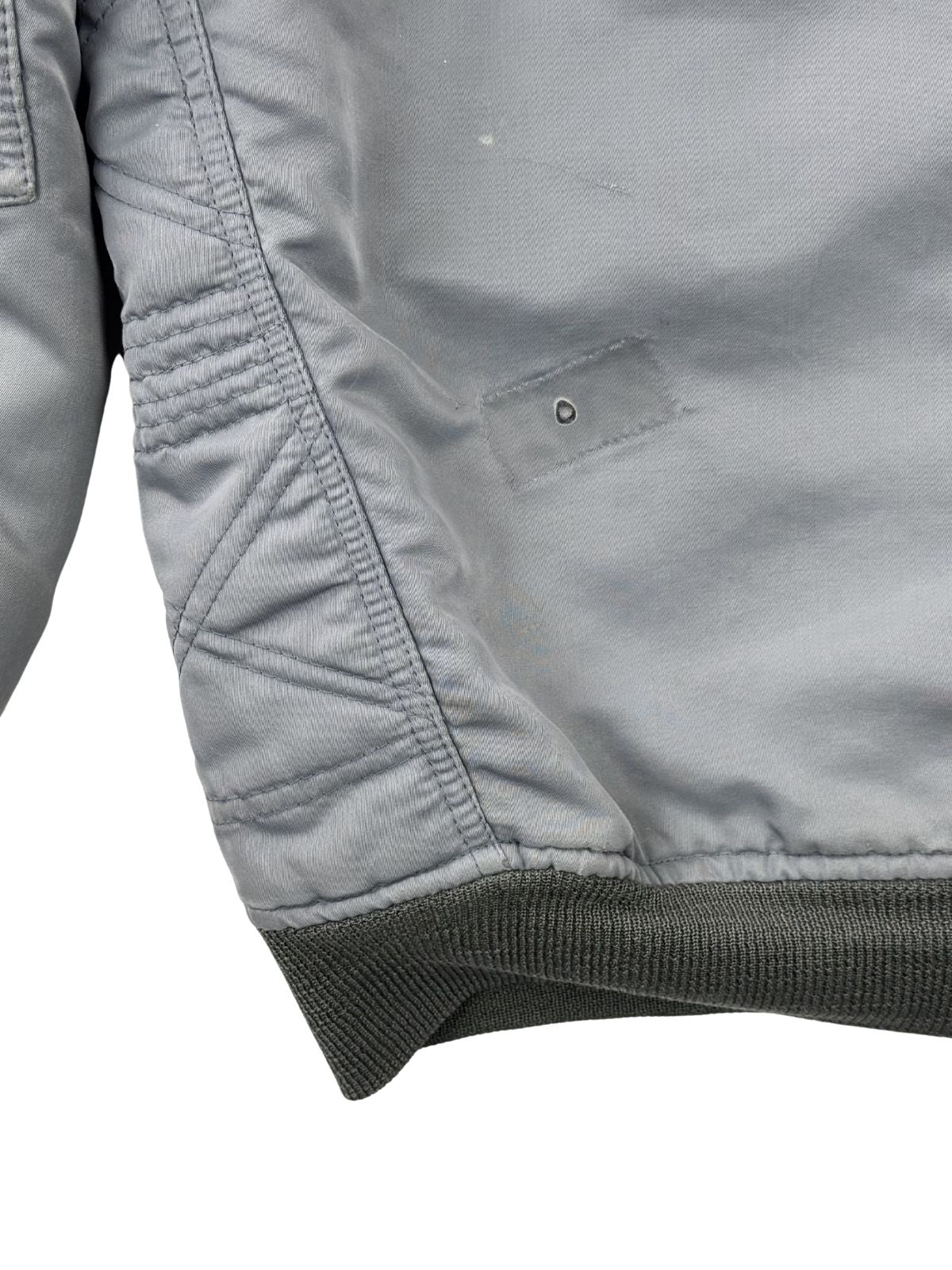 Detailaufnahme des seitlichen Bundes; zeigt die Kombination aus dem grauen Rippstrick-Saum, einem kleinen Loch im Stoff und dem Beginn der gesteppten Diamond-Quilt-Ellbogenverstärkung.