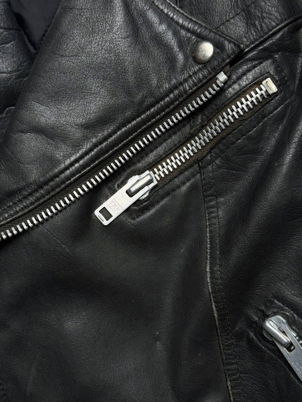Makroaufnahme der schrägen Brusttasche mit silbernem YKK-Metallreißverschluss auf narbigem schwarzem Leder.
