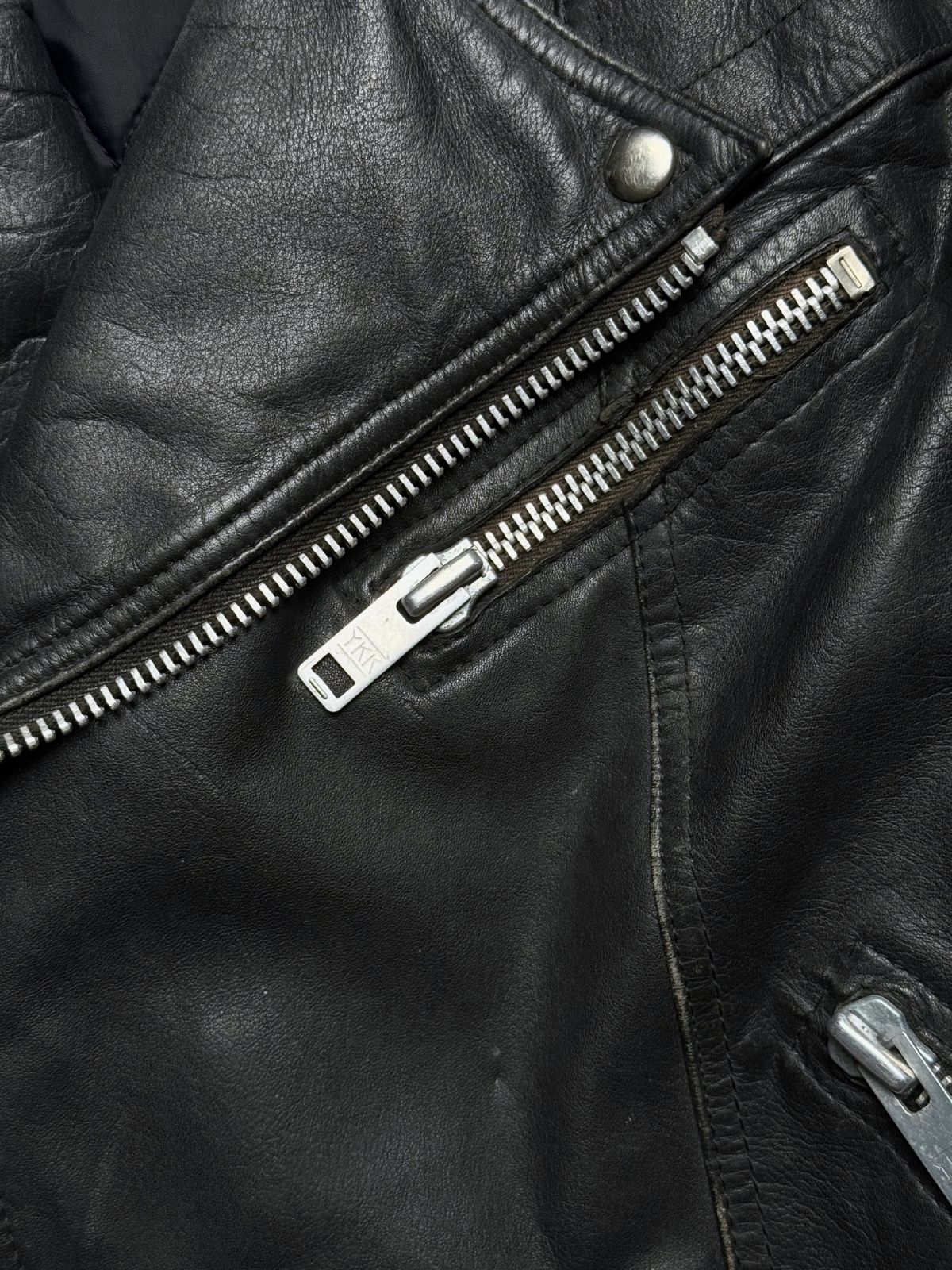 Makroaufnahme der schrägen Brusttasche mit silbernem YKK-Metallreißverschluss auf narbigem schwarzem Leder.