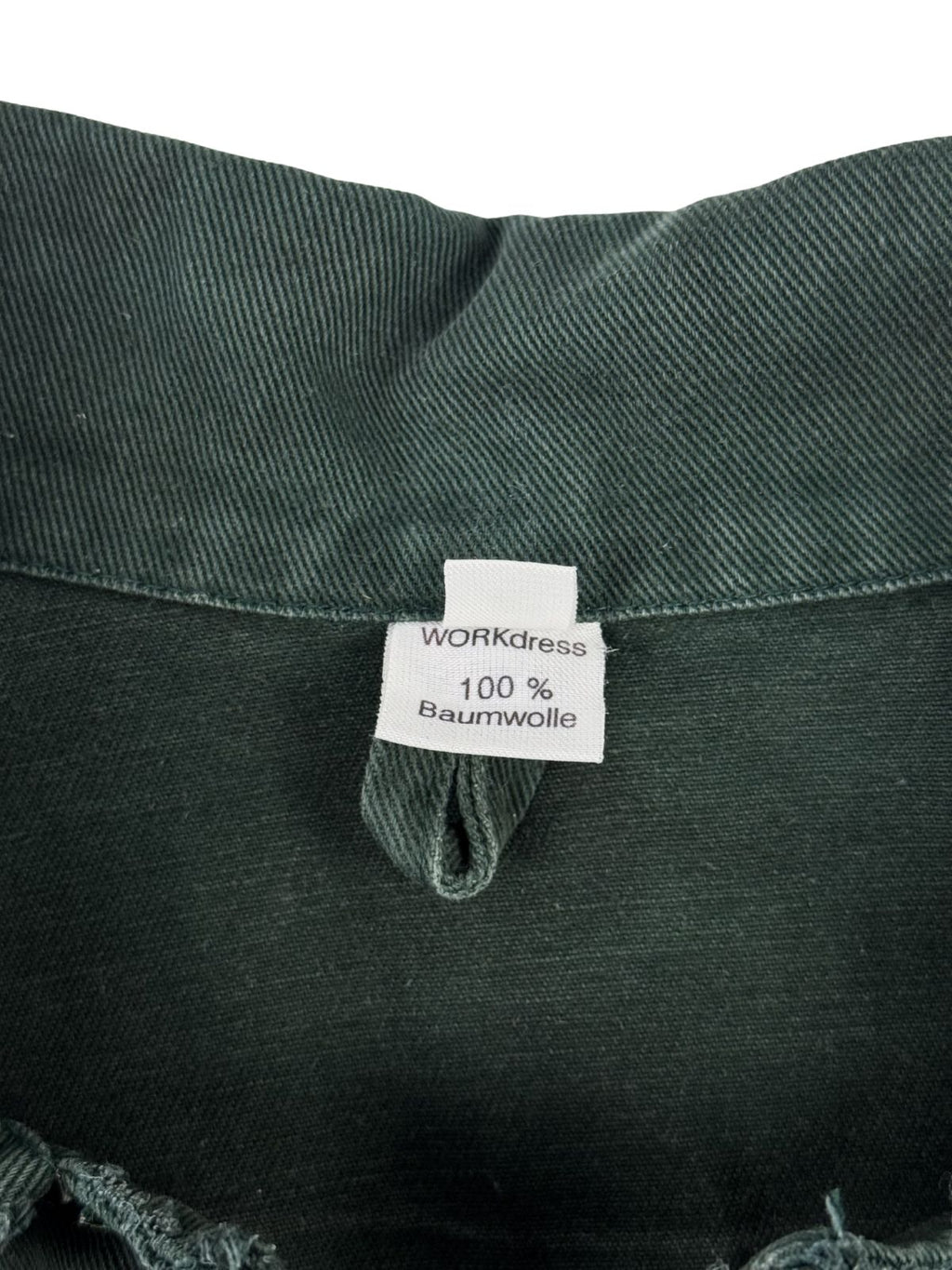 Nahaufnahme des inneren Nackenbereichs mit Blick auf das weiße Stoffetikett der Marke „WORKdress“ mit der Materialangabe „100% Baumwolle“ und einer praktischen Aufhängeschlaufe aus grünem Canvas.
